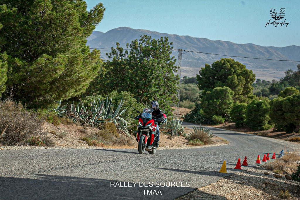 Un pilote de rallye sur sa BMW S1000XR rouge, vêtu d'une veste noire, d'un jean bleu, avec un casque et des bottes noirs, navigue à travers un paysage naturel verdoyant