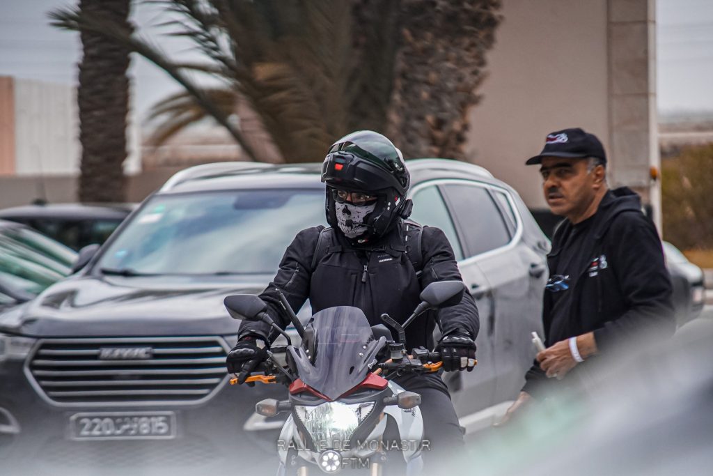 Un pilote de rallye sur sa Honda CB1000R rouge, vêtu d'une tenue noire avec un casque, des gants, et un masque gris et noir, se prépare pour le rallye
