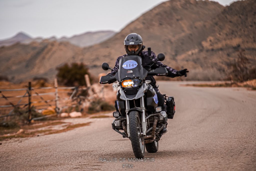 Un pilote de rallye sur sa BMW R1200 GS Adventure noire, en saluant la caméra lors d'un rallye sur route. Sa tenue entièrement noire, y compris son casque, gants et bottes, reflète son engagement dans la compétition