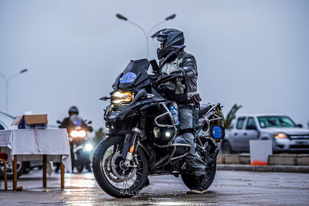 Un pilote de rallye sur sa BMW R1200 GS Adventure, vêtu de noir et gris, se préparant pour une course hivernale, en arrière-plan, un autre pilote de rallye se prépare également pour la compétition